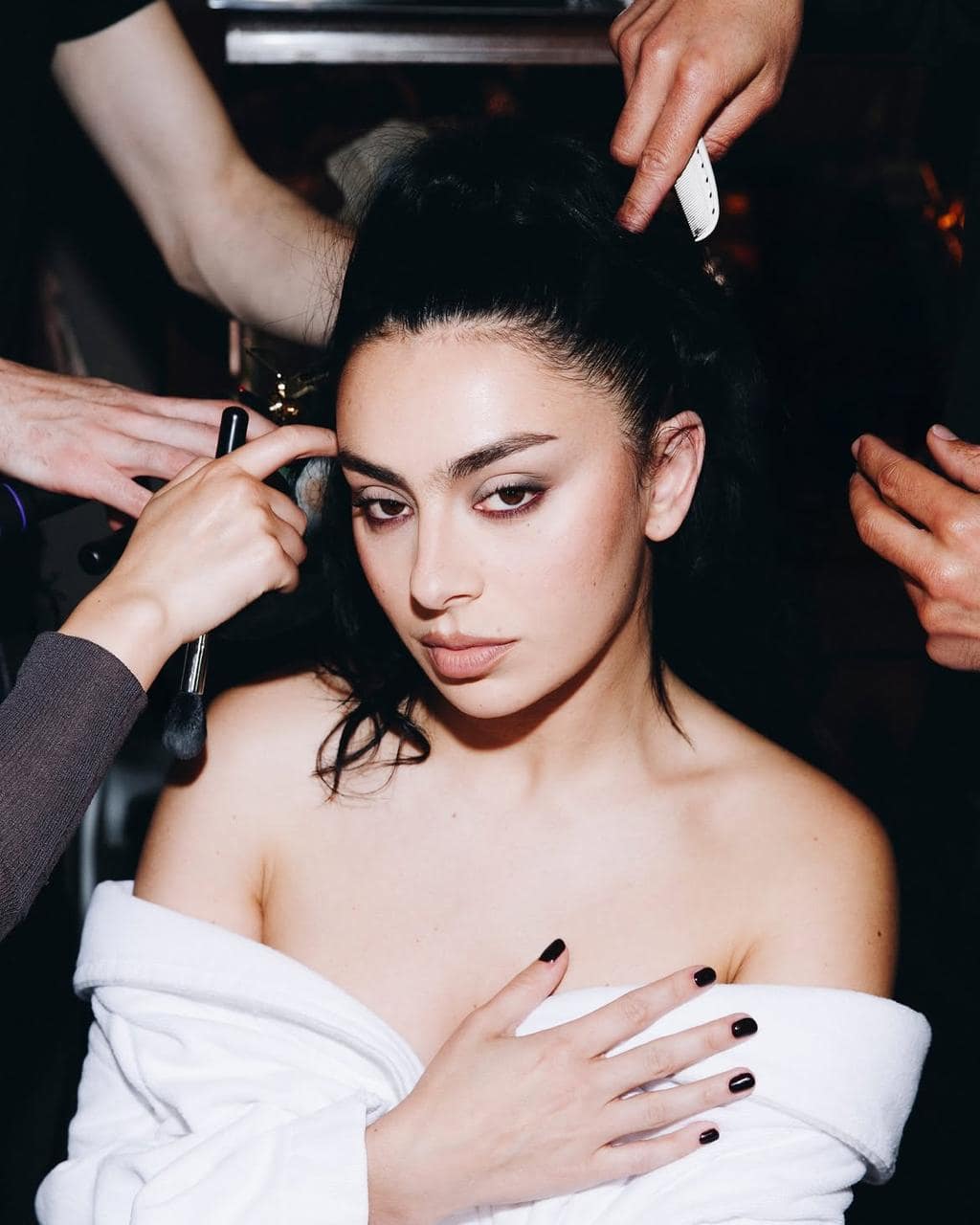 Close-up of singer Charli XCX during makeup application, featuring soft espresso smoky eye shadow, bare lashes, natural fresh skin, and messy black nail polish.