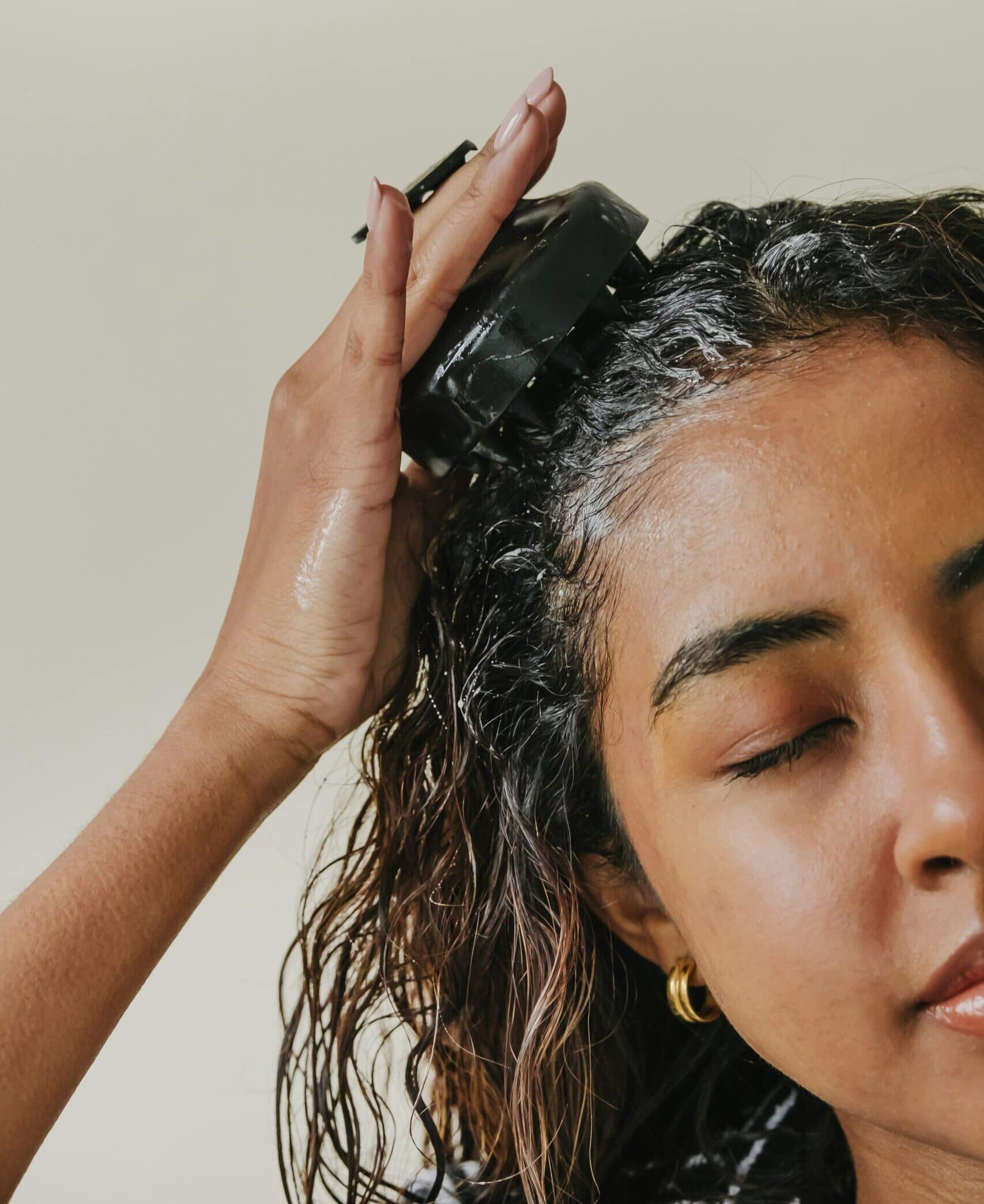 A close-up shot of a woman with her eyes closed, using a black silicone scalp massager to work white shampoo lather into the roots of her damp hair.