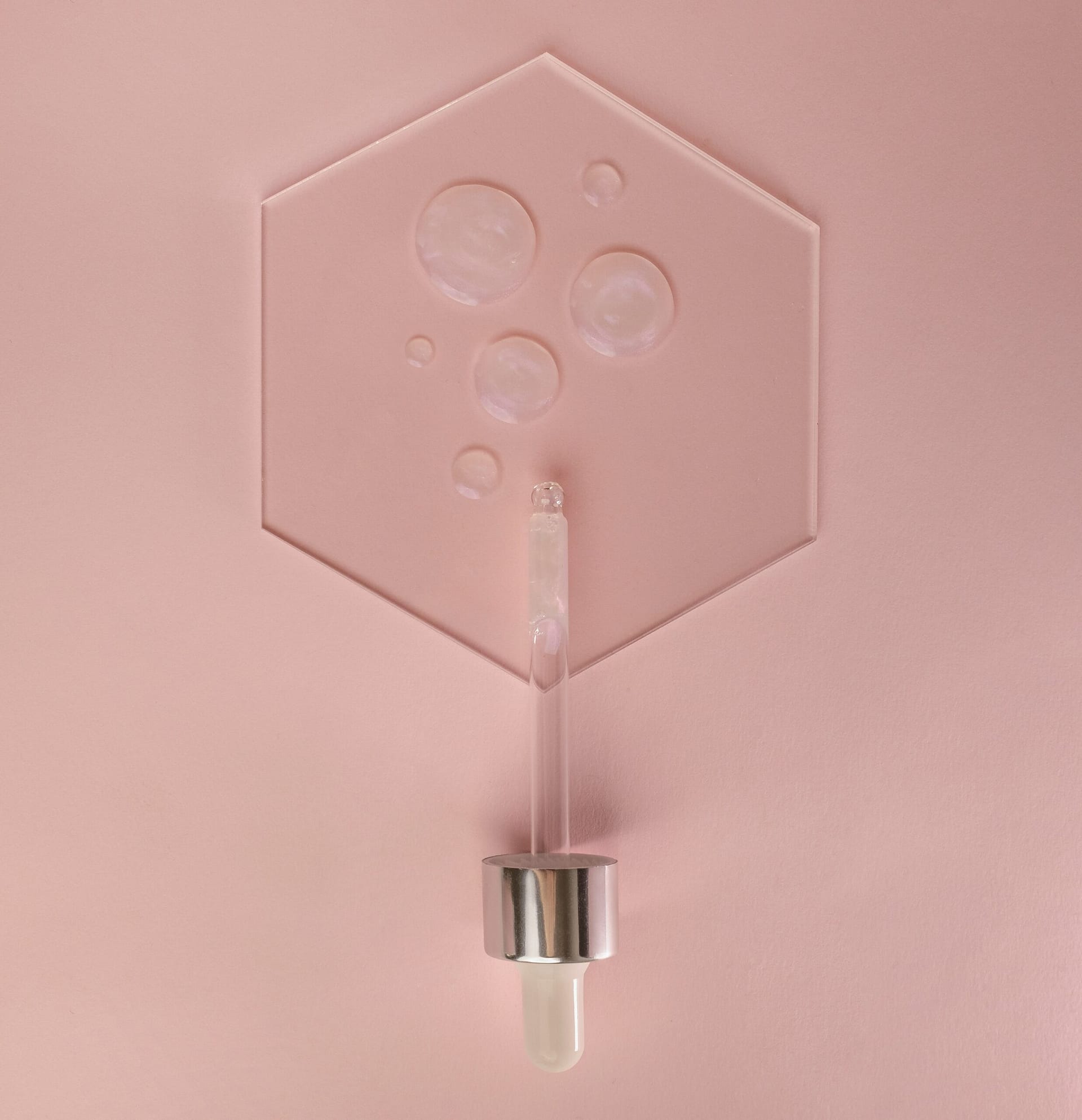 Top view of a glass pipette dropper dispensing a clear liquid serum inside a raised hexagon outline on a minimalist dusty rose background with clean aesthetic textures.