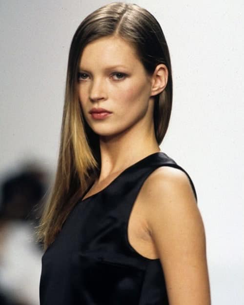 Kate Moss on the Calvin Klein Spring 1994 runway showcasing 90s restrained makeup with satin skin, narrow brows, matte taupe eyeshadow, and a defined brown lip liner outline.