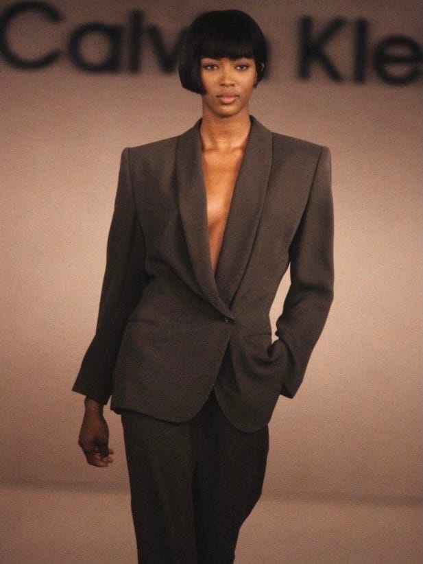 Naomi Campbell walking the Calvin Klein Spring 1990 runway in a grey tailored pantsuit, showcasing a 90s minimalist makeup look with bare eyes and a muted caramel-coffee lip.