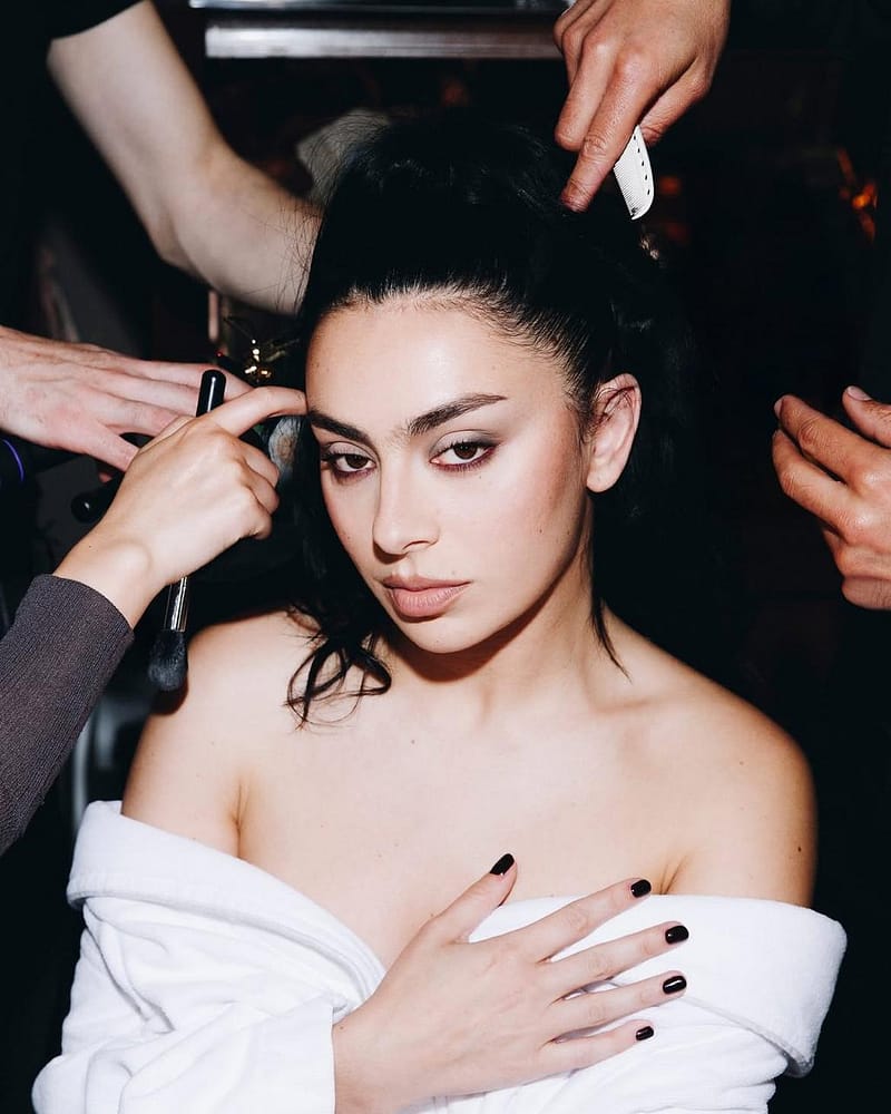 Close-up of singer Charli XCX during makeup application, featuring soft espresso smoky eye shadow, bare lashes, natural fresh skin, and messy black nail polish.