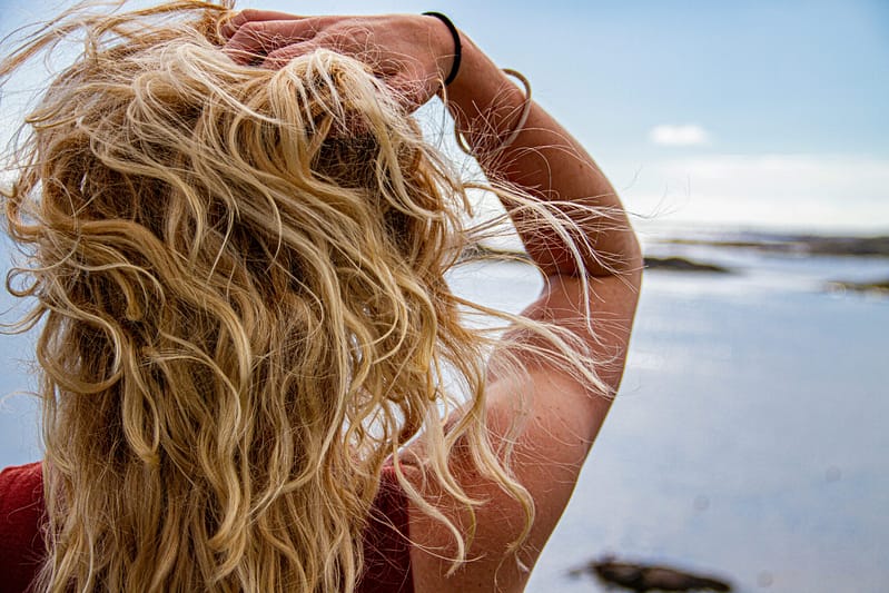 Back view of a woman with naturally textured blonde wavy hair running her hand through her hair against a calm sea background.
