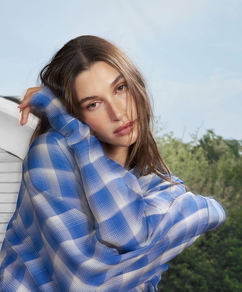 A close-up portrait of Hailey Bieber showcasing the sheer complexion technique. Her skin has a fresh, translucent tinted moisturizer finish with natural skin texture showing through. She is wearing a blue and white checkered shirt, leaning against a white surface outdoors under soft, diffused daylight with green trees and a clear sky in the background.